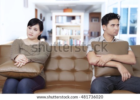 Two young Asian couple having problems at home while sitting on the couch and holding pillow