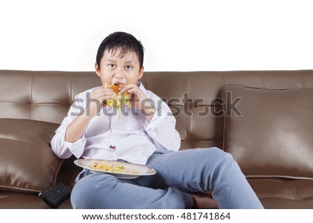 Picture of a little boy enjoying cheeseburger while sitting on the couch and watching tv