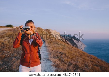 young blonde woman takes the photo on the retro camera on a background of mountain peaks, cliffs and the sea