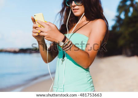 Young pretty brunette woman posing in the beach, listening music in smartphone player, smile, happy face, hipster style, outdoor portrait, reading text, message, sunglasses, close up, sea, Thailand,