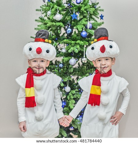 Boys, twins in carnival costumes of snowmen. Stand hold hands before the Christmas fir-tree.