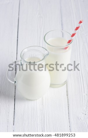Glass of milk with stripped red paper straw and jug of milk on white wooden table still life