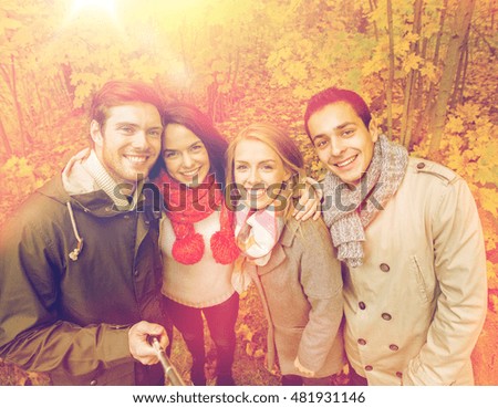 season, people, technology and friendship concept - group of smiling friends with smartphone or digital camera and selfie stick taking picture in autumn park
