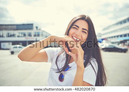 Laughing attractive young woman standing outdoors in an urban environment making a heart gesture with her hands symbolic of love and romance