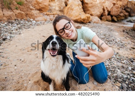 Smiling young cheerful girl in eyeglasses having fun with her dog at the beach and taking selfie with smartphone