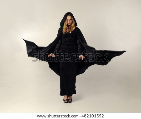 full length portrait of a pretty blonde lady wearing a gothic black dress and hooded cloak.
standing pose against a grey background. Royalty-Free Stock Photo #482101552