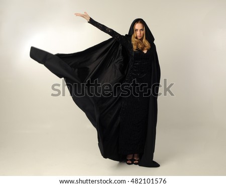 full length portrait of a pretty blonde lady wearing a gothic black dress and hooded cloak.
standing pose against a grey background.