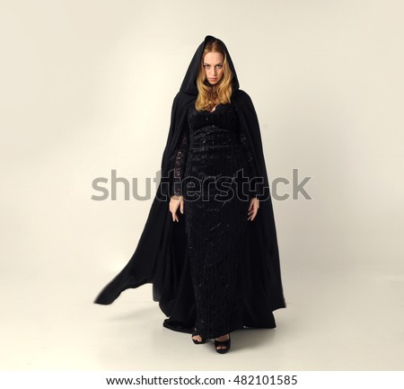 full length portrait of a pretty blonde lady wearing a gothic black dress and hooded cloak.
standing pose against a grey background. Royalty-Free Stock Photo #482101585
