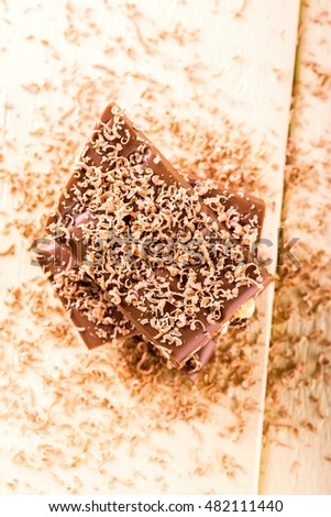 Vertical photo with top view on stack from brown chocolate which contains nuts. Sweet chocolate shavings are spilled everywhere on the picture. Dessert is placed on wooden board with light color.