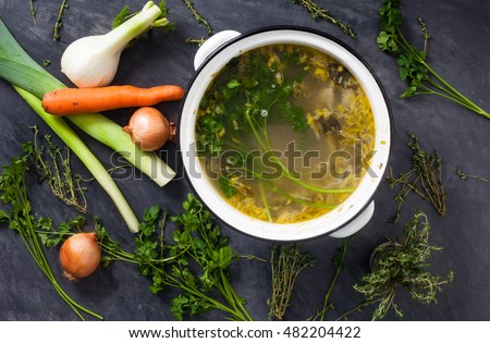 A large pot of homemade coocked fresh organic vegetable stock or broth with fennel, parsley, thyme, onions, carrot, mushrooms and leeks. Stone grey background with vegetables on it. 