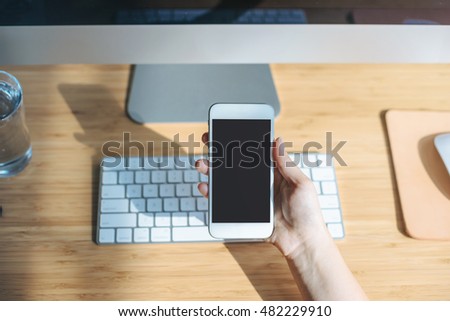 Closeup of female hand using smart phone with blank screen at office, Female hand holding and using modern smartphone with workplace on the background, Workplace in the early morning, Shallow DOF.