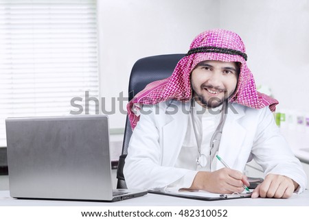 Portrait of Arabian doctor working in the clinic room with clipboard and laptop on the table, smiling at the camera