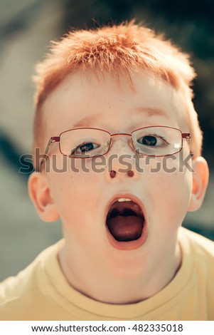 Funny little boy with red hair in eyeglasses with open mouth outdoors on sunny summer day