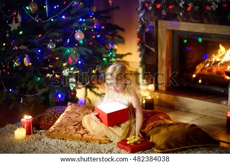 Happy girl opening Christmas gifts by a fireplace in a cozy dark living room on Xmas eve