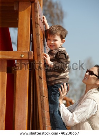Happy mother helping little boy to climb wooden jungle gym on playground, sunlit outdoor.