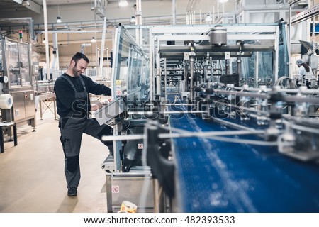 Happy smiled worker doing his job on Robotic factory line for processing and quality control of pure spring water bottled into canisters. Low light and small amount of noise visible. Royalty-Free Stock Photo #482393533