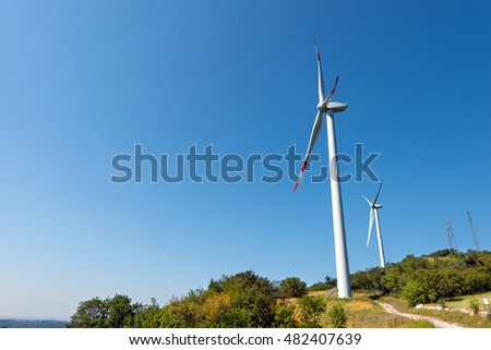 Two white and red wind turbines on a green hill with clear blue sky and power lines. Verona, Italy