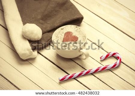 Cup of coffee with heart shape and christmas candy near Santas hat on white wooden background.