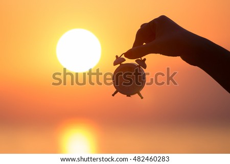 Silhouette of female hand holding small alarm clock and sunrise over sea in the background. Getting up early concept. Royalty-Free Stock Photo #482460283