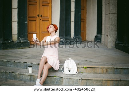 girl doing selfie. woman with dyed red hair in a pale pink dress with a backpack white color drinking coffee, pictures of themselves on their mobile camera phone, sitting on the stairs summer day