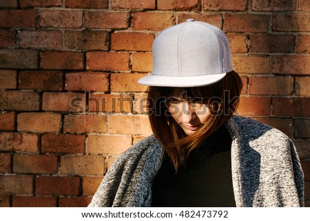Young girl in a blank white cap is standing in a sunlight on a brick background. A female with brown hairs wearing street wear is looking down while standing beside the brick wall.