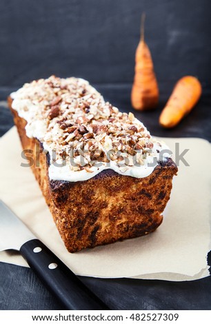 Delicious homemade carrot cake with sweet butter cream icing and finely chopped nuts. A wonderful dessert on the baking paper with a knife nearby and two blurred carrots behind on black background.