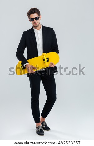 Full length portrait of a young handsome businessman in suit and suglasses holding yellow skateboard over gray background