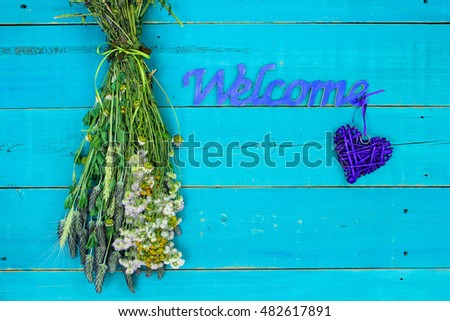 Welcome sign with heart by bouquet of dried meadow flowers hanging by rope on antique rustic teal blue wood background; Mother's Day background with copy space