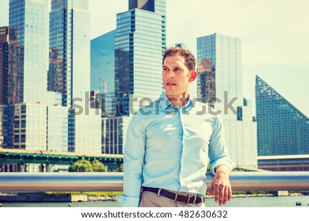 American Man summer vacation in New York. A young guy wearing white shirt, standing by metal fence in busy business district with high buildings under sun, looking away, thinking. Filtered look.