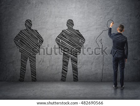 Back view of a businessman drawing dark men's silhouettes on concrete wall. Employment issues. Outlines and contours. Royalty-Free Stock Photo #482690566