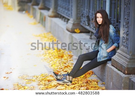 Young pensive brunette girl sitting on a concrete curb around a beautiful iron fence, under the feet of yellow autumn maple leaves.