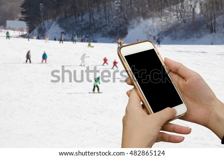 woman hands holding mobile smart phone over blurred skiers in the ski field, winter background