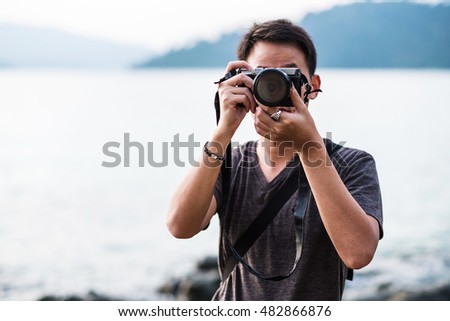 Asian guy traveler taking with vintage photo camera at tropical beach, selective focus on camera