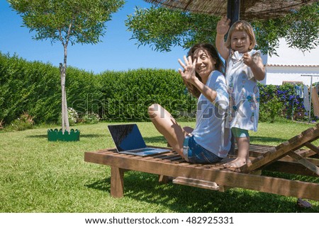 brunette woman with white shirt and jeans shorts with laptop blank screen and two years old blonde child on lounge deck chair at garden saluting hands up