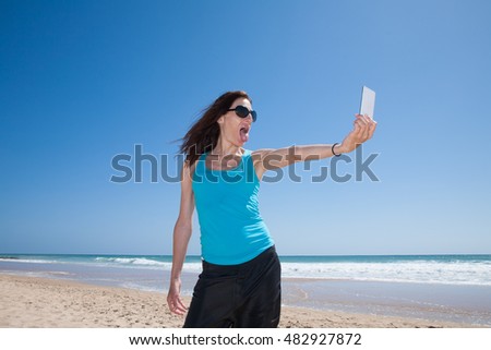 portrait of brunette woman with black sunglasses and blue cyan shirt teasing sticking out tongue making selfie with mobile phone smartphone at beach with sea behind