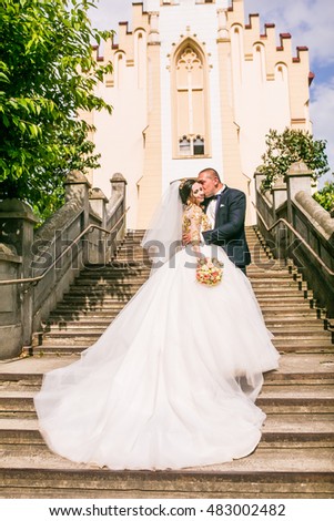 bride and groom walking near trees and church. Woman in yellow gold crown. Decorated yellow beads dress. Embracing couple. Looking each other. Adorable girl. Emotional picture.