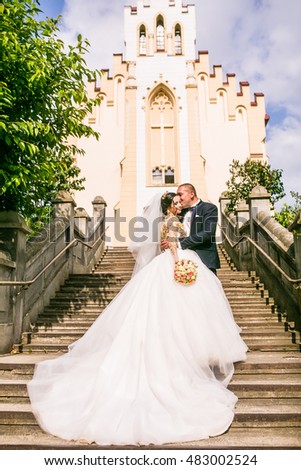 bride and groom walking near trees and church. Woman in yellow gold crown. Decorated yellow beads dress. Embracing couple. Looking each other. Adorable girl. Emotional picture.