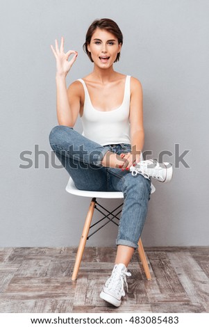 Attractive brunette woman sitting on the chair and showing okay sign over gray background