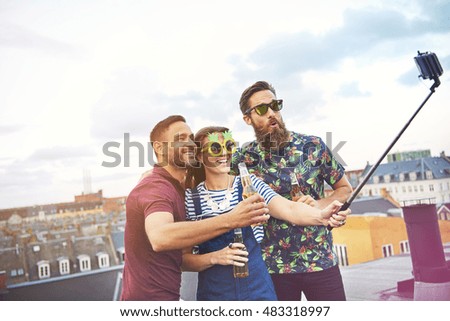 Three happy friends with selfie stick taking a picture of themselves while drinking beers on top of roof in city