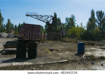 An old rusty crane for lifting cargo in an industrial area against a ruined building, an old asphalt pavement and a blue sky. Abandoned and stolen industrial base. Destruction and vandalism.