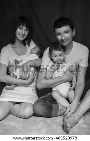Little infant baby boy sleeping laying on mothers arms, cute 2 years girl laying on fathers arms smiling. Neutral black background, black and white picture. All in white clothes. Happy family