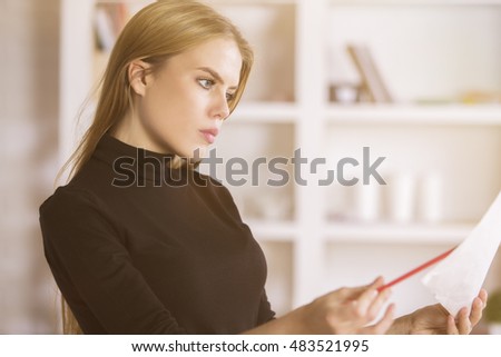 Portrait of gorgeous european businesslady doing paperwork in modern office. Blurry shelves with items in the background