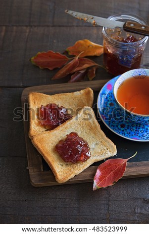 Instagram looking picture of cup of tea, two toasts, jar of jam and red leaves over rustic wooden background. Color toning, lifted shadows