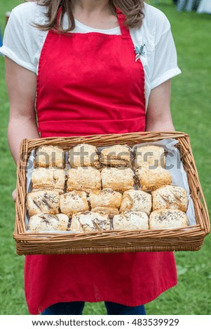woman in red apron cropped from neck down holding basket of homemade savoury pastries and sausage rolls with poppy and sesame seeds