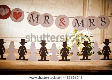 Bride and Groom Paper Doll Cut Outs Lined Up on Rustic Wooden Table in front of White Rose Bouquet and Underneath Mr and Mrs Banner Hanging Up at Wedding Celebration or Bridal Shower