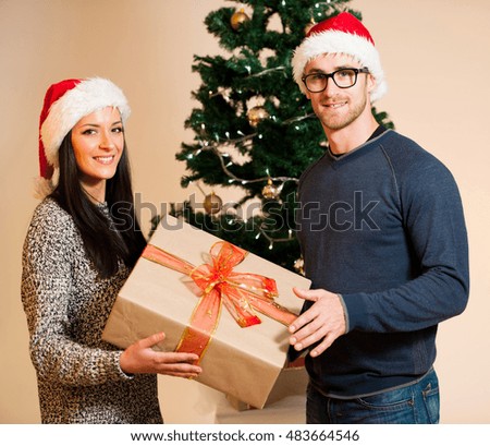 A young couple with santa hat standing in front of the Christmas tree and giving gift to each other.