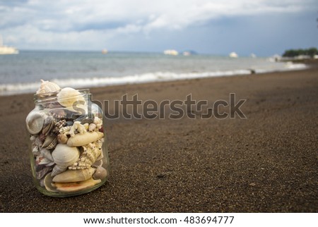 Seashells in jar on the sand beach. Tropical landscape with sea and white boats. Shells collection from the seashore on seascape background. Vacation memories poster or background template image