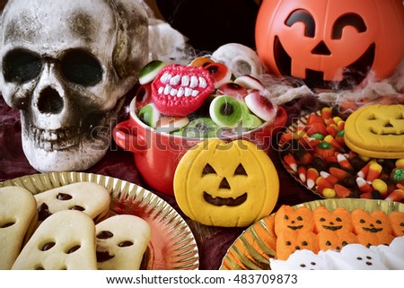 different Halloween candies and cookies on a table decorated with some scary ornaments, such as some skulls and cobwebs and a carved pumpkin