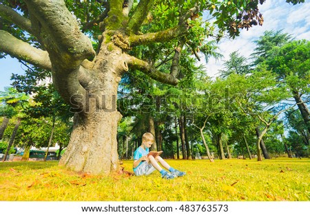 Small boy sitting under big tree on city park and reading book.