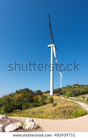 Two white and red wind turbines on a green hill with clear blue sky and power lines. Verona, Italy